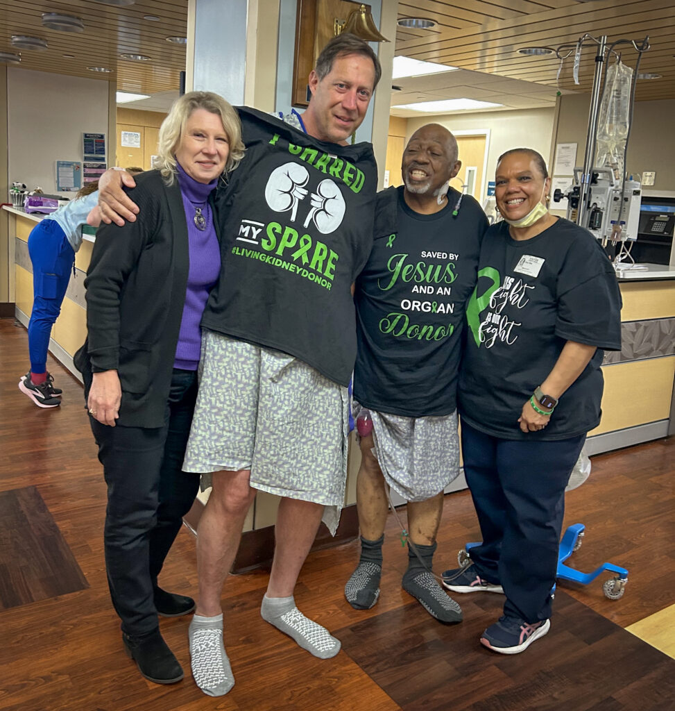 Four people stand smiling in a hospital hallway, two women and two men. The men are both in hospital gowns