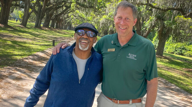 Two men, one Caucasian and the other African American, stand with their arms around each other on a gravel road under a long line of oak trees
