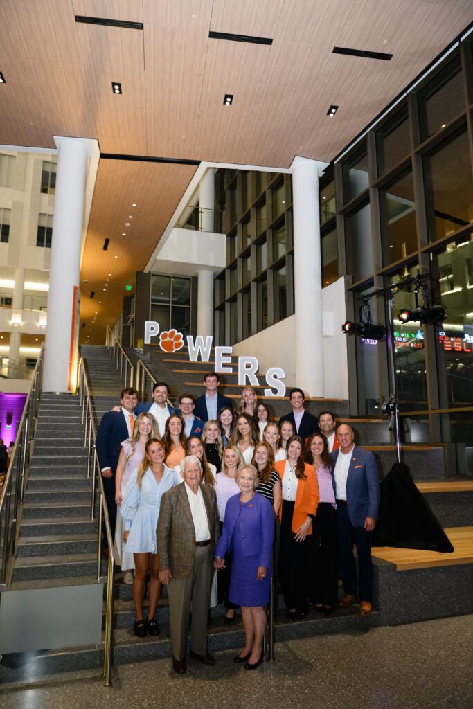 A group of students stand with an older couple on a set of stairs and letters spelling "Powers" are in the background, with an orange Tiger Paw instead of the "O" in Powers.