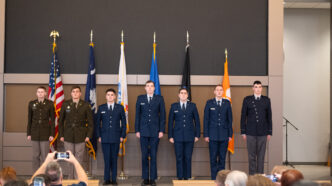 Seven young men standing on a stage, wearing uniforms, six flags mounted behind them.