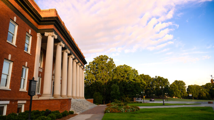 The front columns of Sikes Hall with a cloudy sunset and Bowman field in the background.