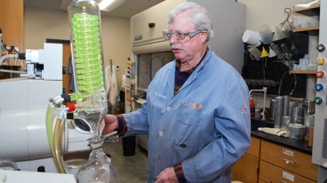 A man wearing a blue lab coat and safety goggles looks at a machine that has a green coil in a science lab.
