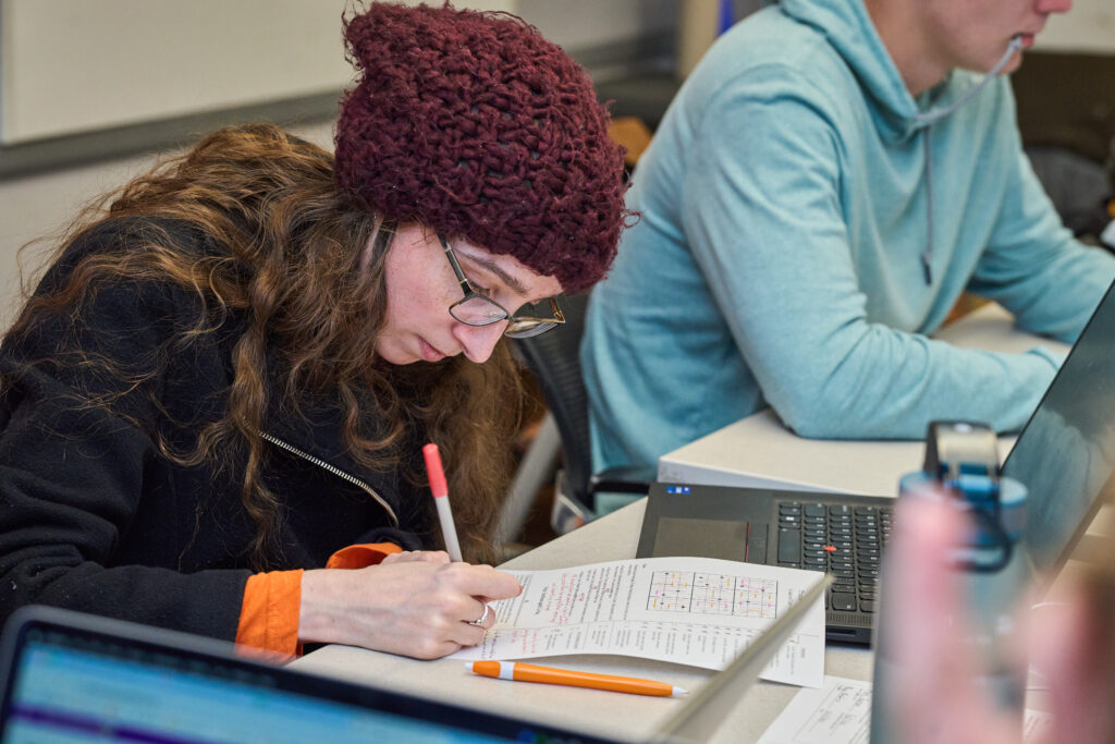 A woman wearing a beanie hat works on a sudoku puzzle at her desk.