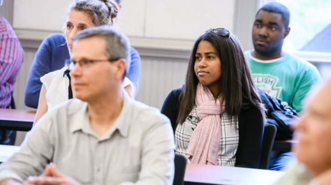A diverse group of adults seated at desks in a classroom or meeting room, listening attentively. In the foreground, a man with glasses sits with hands clasped, while behind him a woman wearing a pink scarf looks forward with a thoughtful expression. Other attendees are visible in the background, all focused on the discussion.