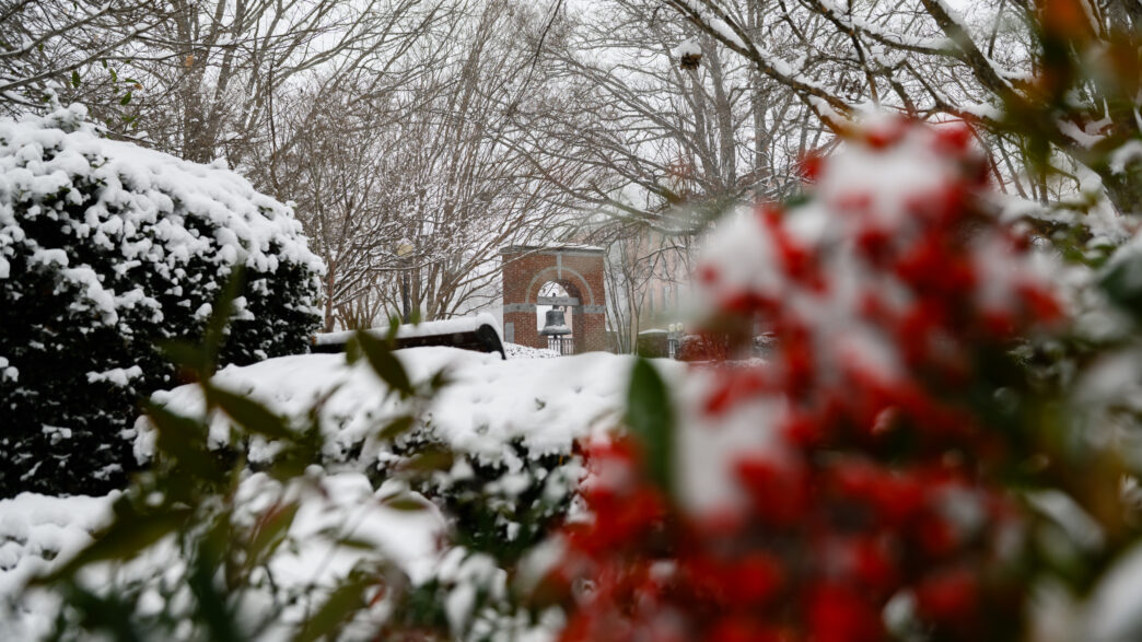 Snow falls on the Carillon Garden bell. Red berries are blurry but visible in the foreground.