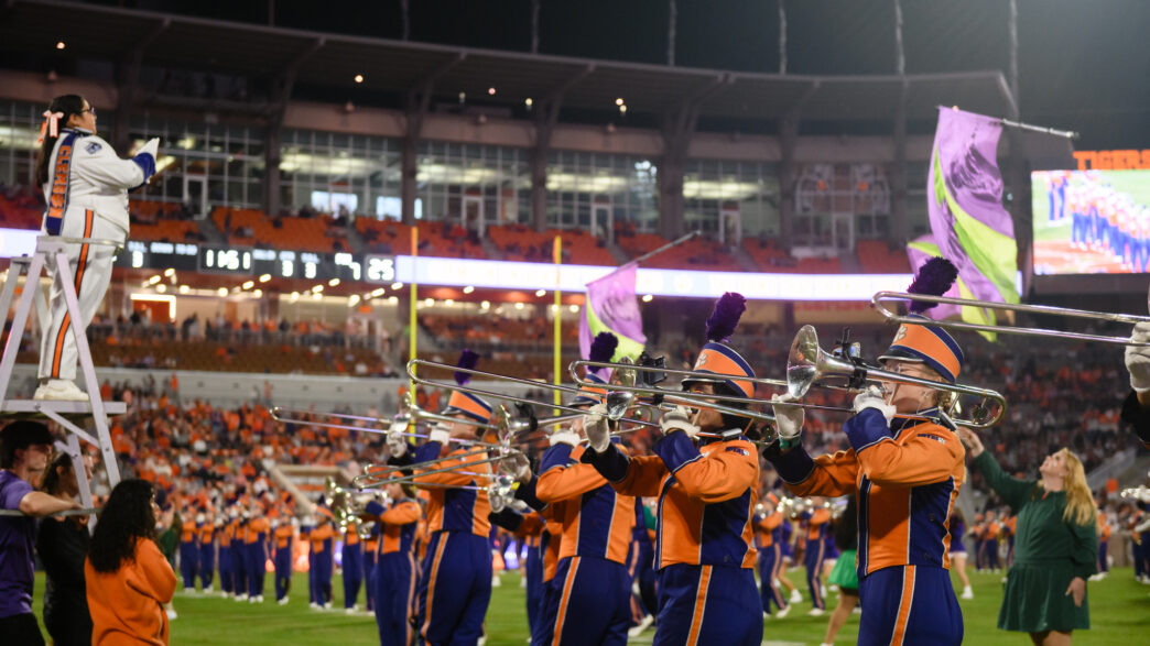 Clemson University Tiger Band trombone line performs on the football field during a night game, with a drum major conducting from a ladder and color guard flags waving in a packed stadium.