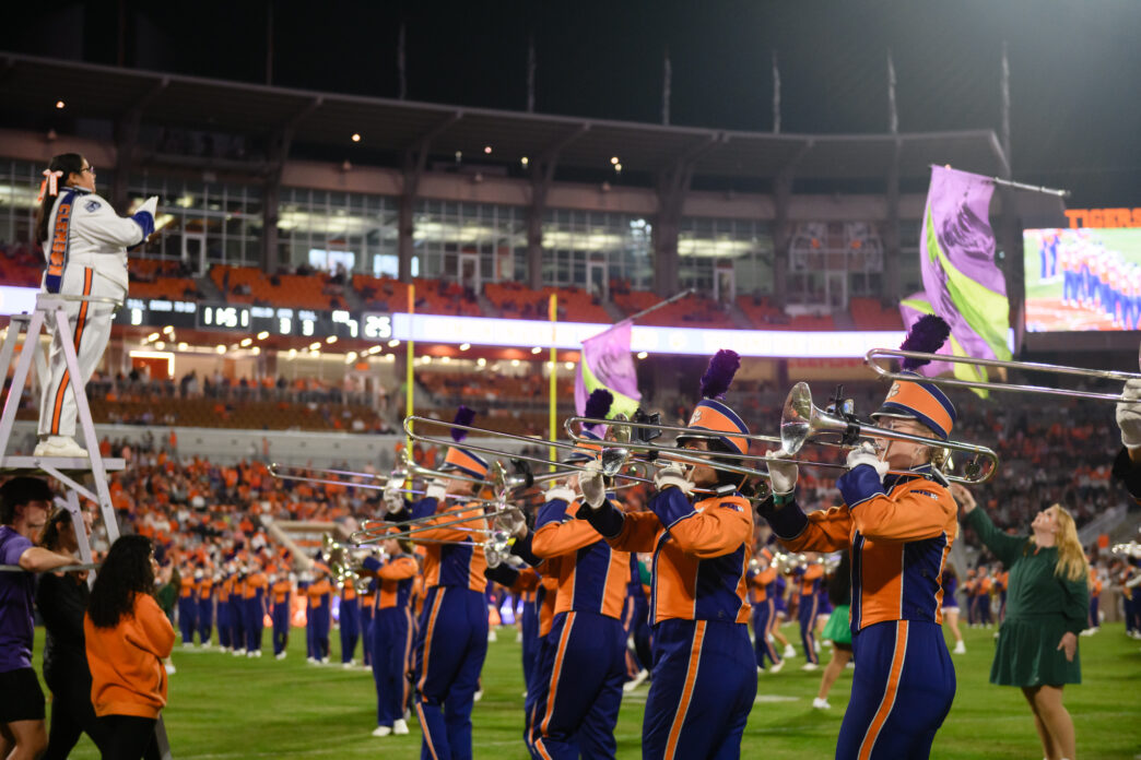 Clemson University Tiger Band trombone line performs on the football field during a night game, with a drum major conducting from a ladder and color guard flags waving in a packed stadium.