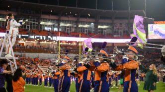 Clemson University Tiger Band trombone line performs on the football field during a night game, with a drum major conducting from a ladder and color guard flags waving in a packed stadium.