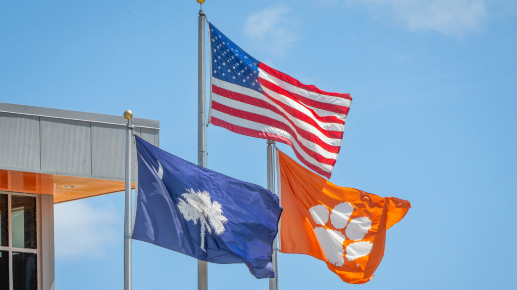 An American flag flies between a South Carolina state flag and a Clemson flag against a blue sky.