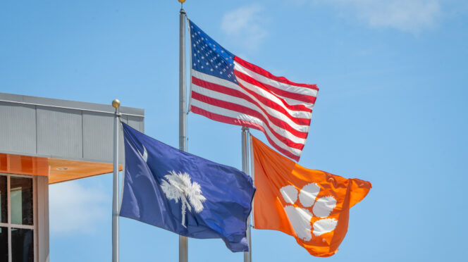 An American flag flies between a South Carolina state flag and a Clemson flag against a blue sky.