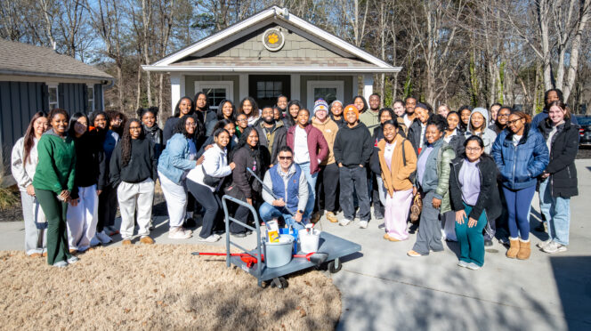 A large group of young people stand and kneel in front of a small house, a cart full of cleaning supplies in front of them.