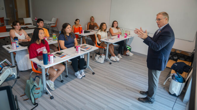 A man in a suit speaks in front of students within a classroom