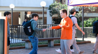 A male in an orange sweatshirt hands a piece of paper to a student in jeans wearing a backpack