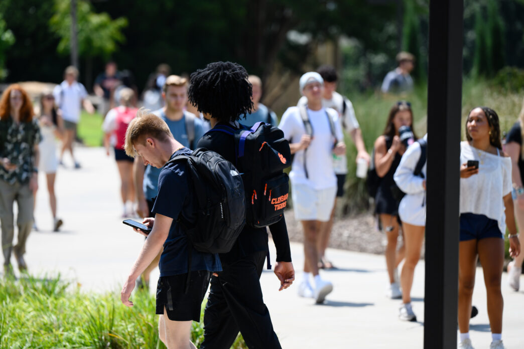 A group of students walking on a sidewalk. One student is looking down at his phone while walking. Another student is walking away from the camera, and he is wearing a backpack. A large group of students walk towards the camera.