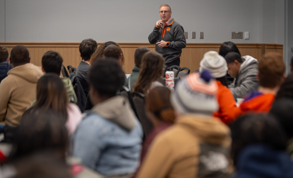 A man in a Clemson-branded jacket holds a microphone up to his mouth and speaks to a large group of people sitting at tables in front of him.