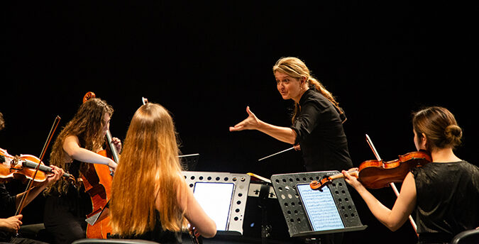A conductor gestures expressively while leading a small string ensemble—violinists and a cellist—during a live performance on a darkened stage, with musicians reading from music stands.