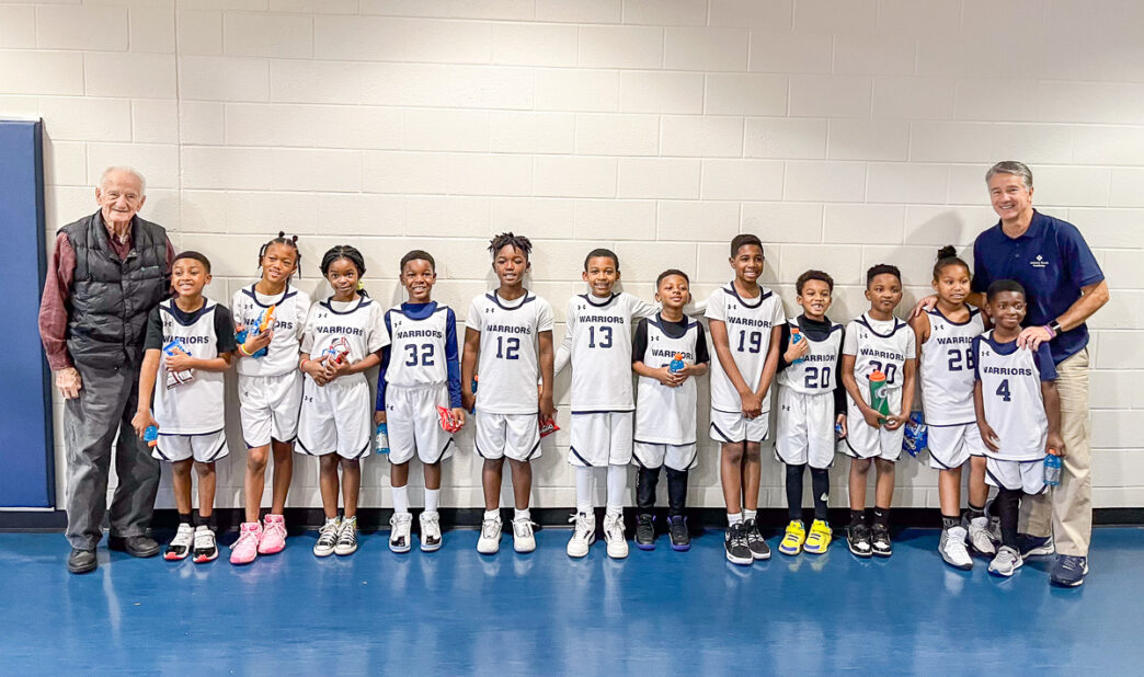 A youth basketball team lined up against a wall of a gym with their two coaches, the one on the right is Tony Perticone, a Clemson alumni who started the basketball program at Atlanta Youth Academy.