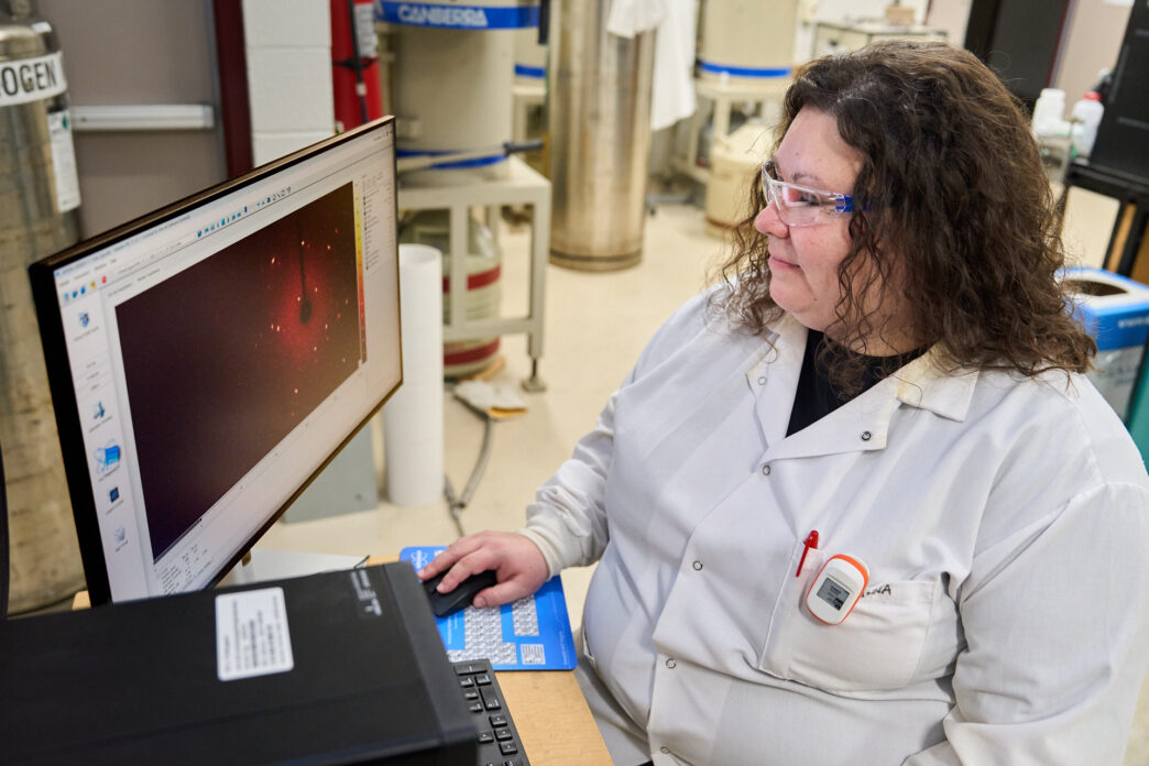 A woman wearing a white lab coat and glasses looks at a computer screen while sitting at a desk in a science lab at Clemson University.