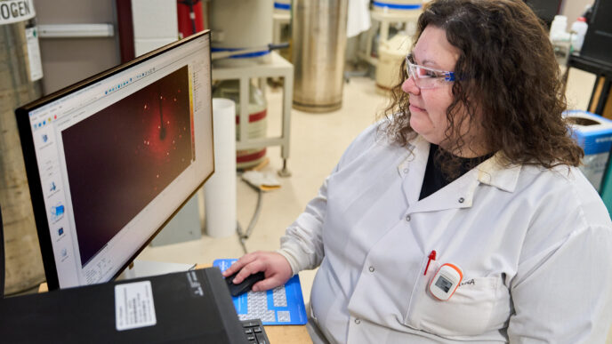 A woman wearing a white lab coat and glasses looks at a computer screen while sitting at a desk in a science lab at Clemson University.