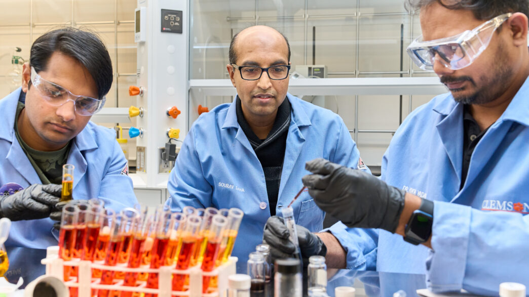 Two men wearing blue lab coats, gloves and goggles are sitting at a ltable in a science lab working with test tubes while another man watches.