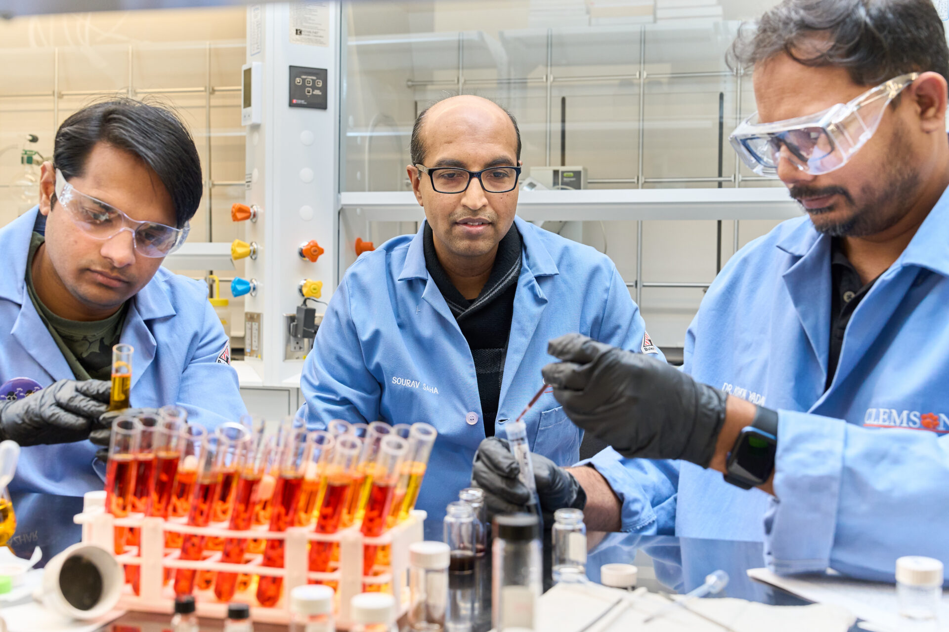 Two men wearing blue lab coats, gloves and goggles are sitting at a ltable in a science lab working with test tubes while another man watches.