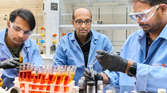 Two men wearing blue lab coats, gloves and goggles are sitting at a ltable in a science lab working with test tubes while another man watches.