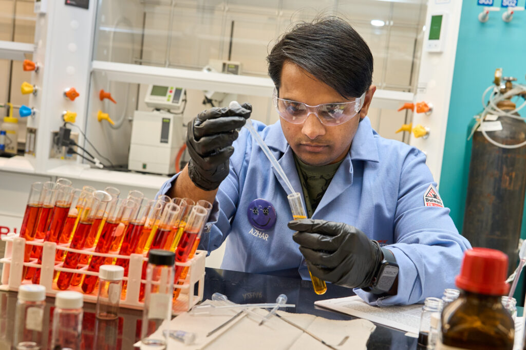 A man wearing a blue lab coat, glasses and gloves puts liquid into a test tube in a science lab.