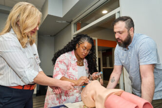A woman tries to intubate a dummy at a demonstration while two people watch.