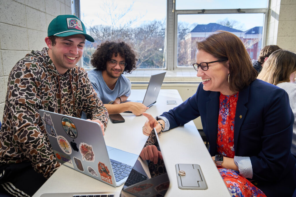 एक आदमी कंप्यूटर की ओर इशारा करता है जबकि दो आदमी देखते हैं।