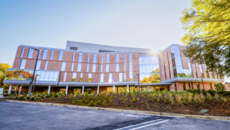The sun sets behind a modern brick building with large glass windows, sending rays of light flaring down across the facade in the shape of a star.