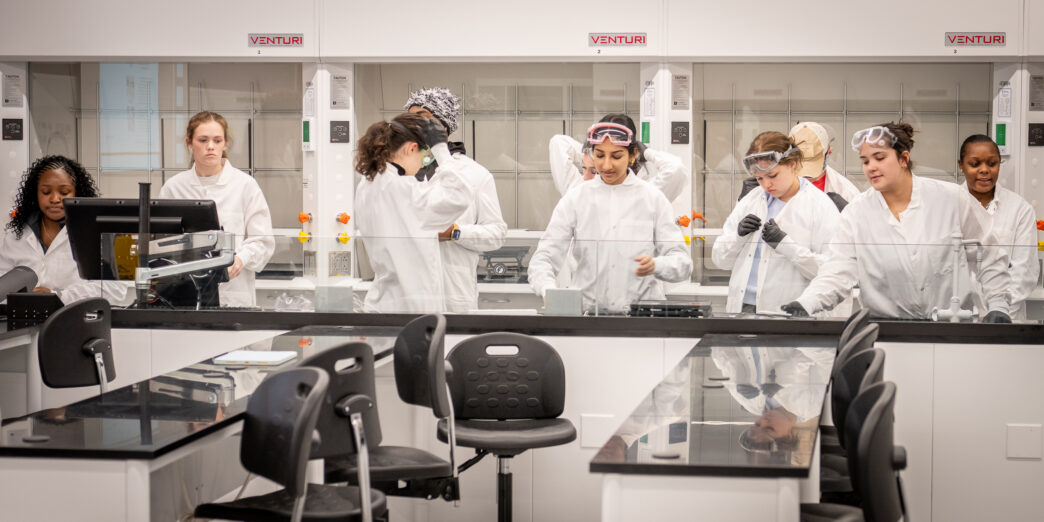 Ten students stand behind a shiny black counter, putting on white lab coats and gloves.