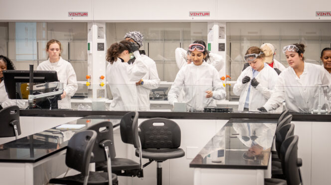 Ten students stand behind a shiny black counter, putting on white lab coats and gloves.