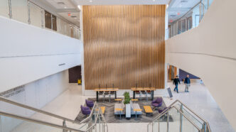 A staircase descends into a large lobby with modern chairs and tables, and two-story wall of 2" notched wood beams lined up in vertical rows.