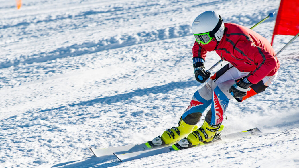 A competitive snow skier wears a red jacket and white helmet as they accelerate down a snowy ski slope with a red flag in the background.