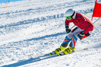 A competitive snow skier wears a red jacket and white helmet as they accelerate down a snowy ski slope with a red flag in the background.