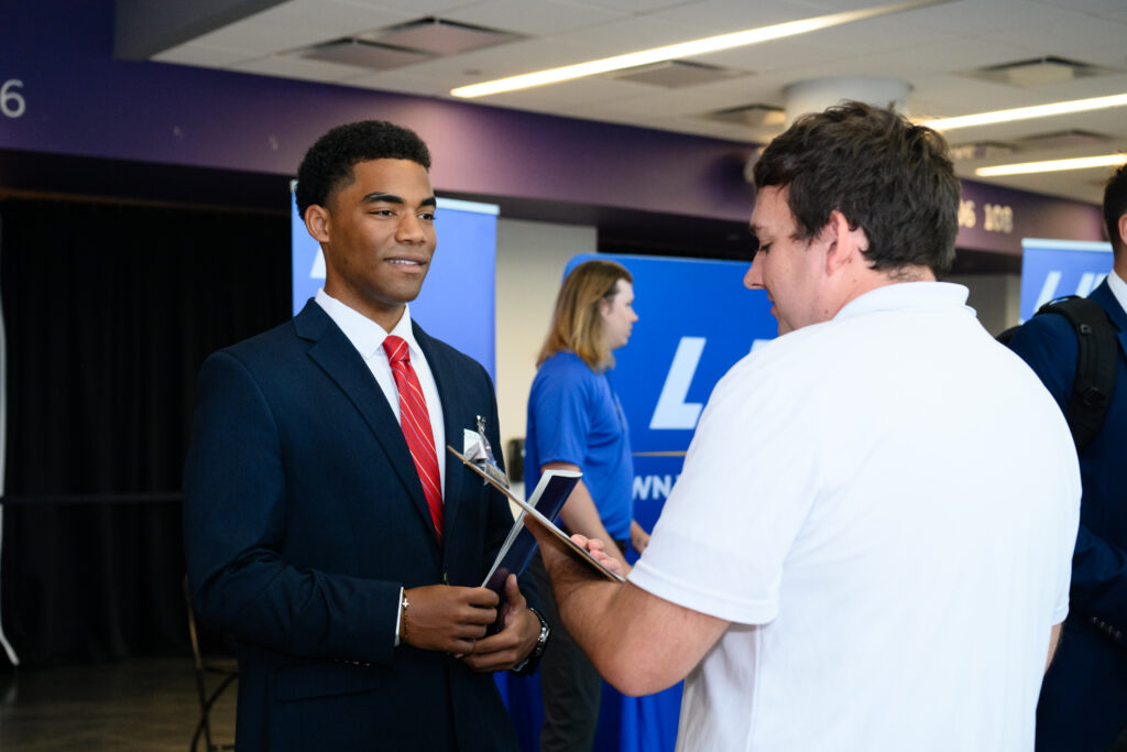 A male in a navy suit with a red tie speaks to another male individual