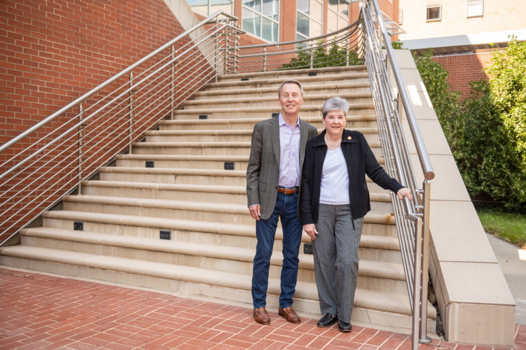 A man wearing a suit jacket, button shirt and jeans and a woman wearing a black jacket, white shirt and dress pants stand at the bottom of an outdoor staircase.