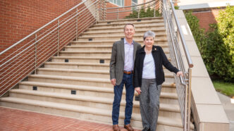 A man wearing a suit jacket, button shirt and jeans and a woman wearing a black jacket, white shirt and dress pants stand at the bottom of an outdoor staircase.