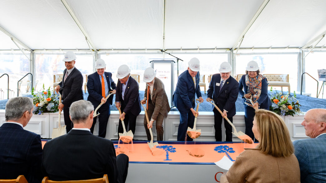 Individuals in suits dig shovels into sand for a ceremonial groundbreaking
