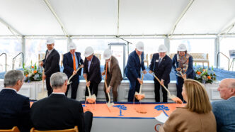 Individuals in suits dig shovels into sand for a ceremonial groundbreaking