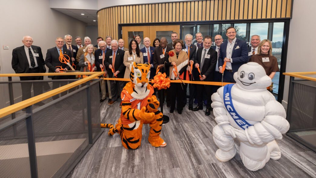 A group of people, several are holding giant scissors, are inside a building, cutting a ribbon for a groundbreaking. The Tiger mascot and Michelin Man mascot are kneeling in front of the group and ribbon.