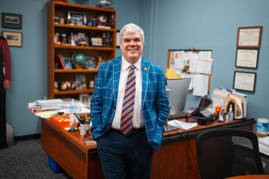 A man in a blue suit stands in front of an office desk