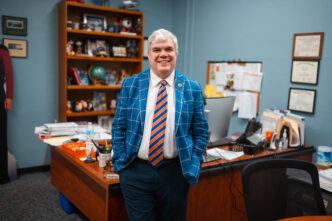 A man in a blue suit stands in front of an office desk
