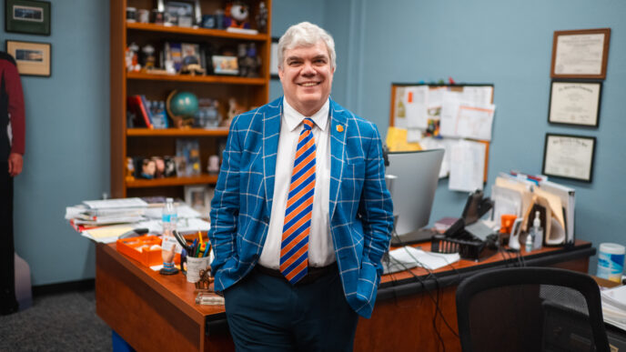 A man in a blue suit stands in front of an office desk