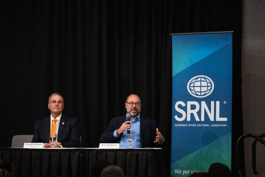 Two men sitting at a table on stage, and one is holding a microphone.