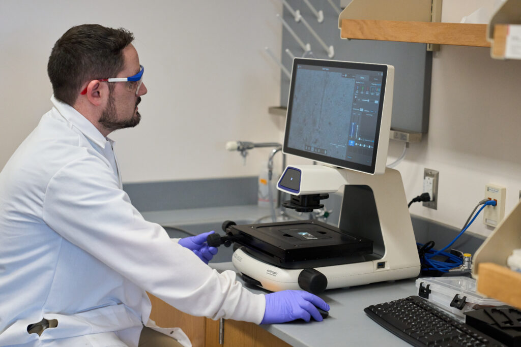 A man wearing a white lab coat, goggles and gloves, looks at a screen of a microscope where he was looking at something on a slide