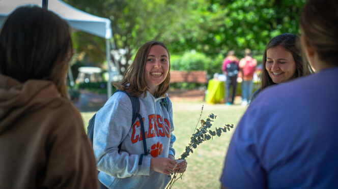 A smiling student wearing a Clemson sweatshirt and backpack talks with three others at an outdoor campus event, holding a small leafy branch, with tents and students visible in the background.