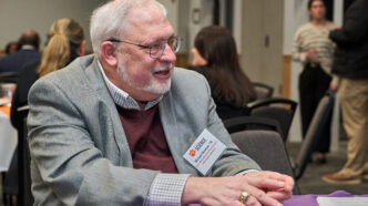 A man wearing a gray suit coat and a burgundy sweater over a white shirt talks with somebody who is sitting at the same table but the other person is not pictured