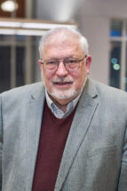 Headshot of Michael Newman, a man with gray hair and a gray mustache and beard wearing a gray suit jacket and burgundy sweater.