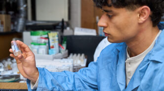 A man wearing a blue lab coat holds up a test tube with crystals in it while in a science lab.